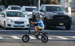 Boy wearing helmet travels across street on electric bicycle.