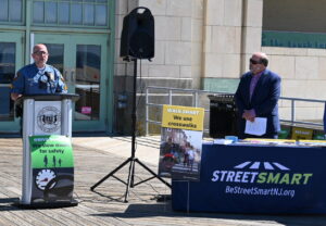 Asbury Park Police Sgt. Michael Casey speaks at podium set up on boardwalk as NJTPA Executive Director David Behrend looks on.